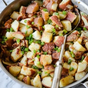 red potatoes, green onions, and crispy bacon in a stainless steel skillet with a large silver serving spoon on a marble surface