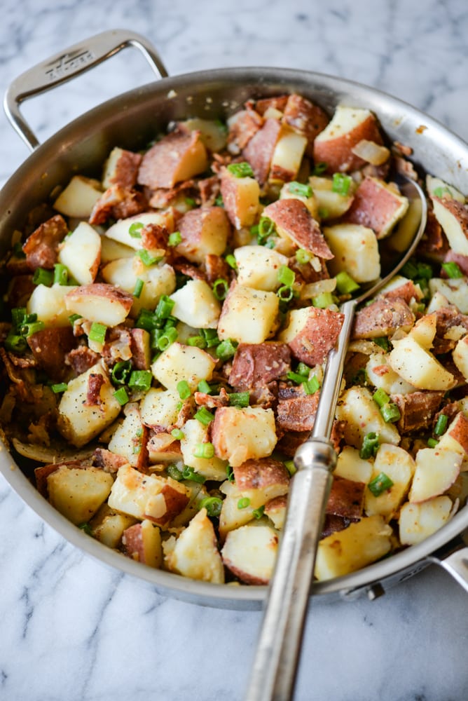 red potatoes, green onions, and crispy bacon in a stainless steel skillet with a large silver serving spoon on a marble surface
