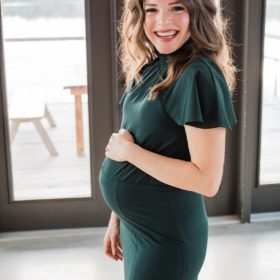 a smiling pregnant woman with long dark wavy hair in a long dark green dress cradles her pregnant belly