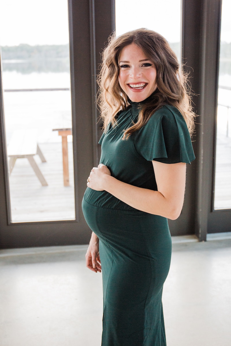 a smiling pregnant woman with long dark wavy hair in a long dark green dress cradles her pregnant belly