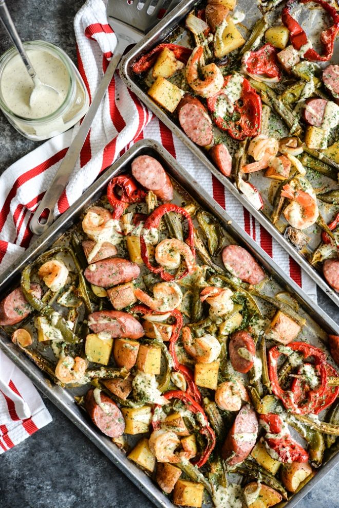 overhead view of a sheet pan loaded with sausage, shrimp, potatoes, asparagus, and red bell peppers with a creamy sauce drizzled over it on top of a dark grey table and red striped napkin