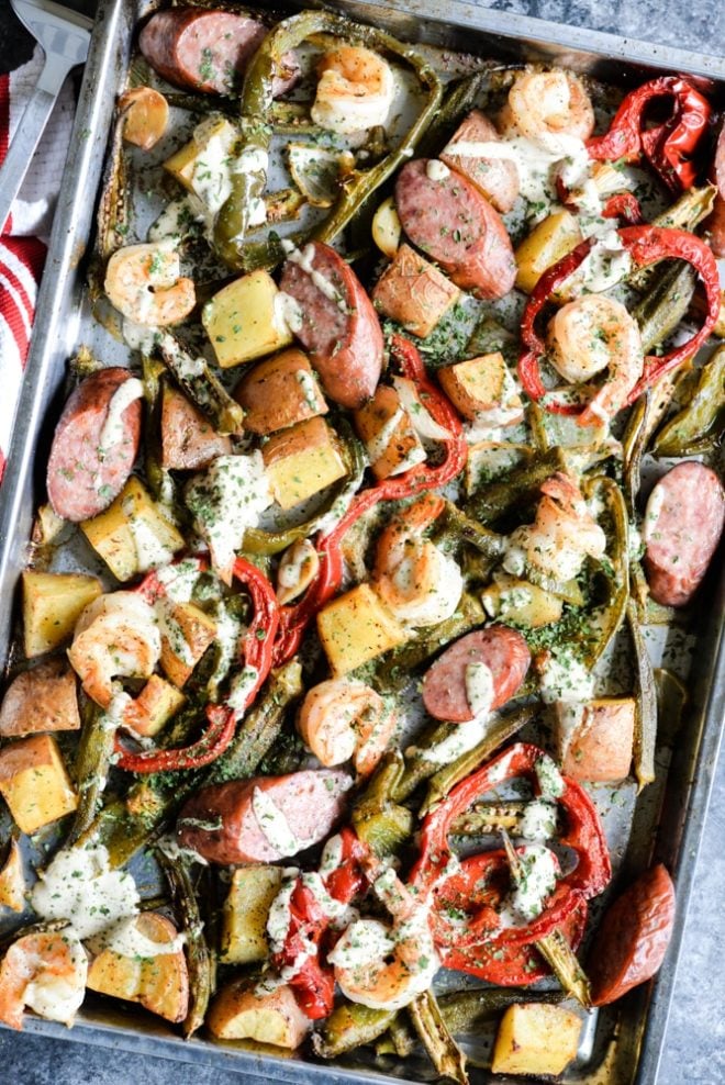 overhead view of a sheet pan loaded with sausage, shrimp, potatoes, asparagus, and red bell peppers with a creamy sauce drizzled over it