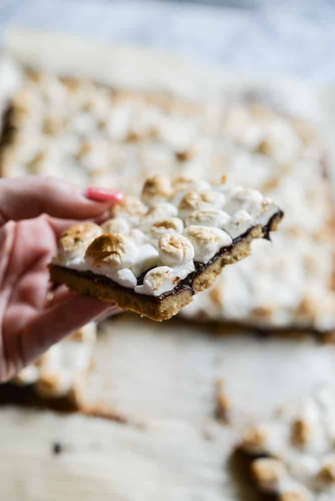 woman's hand with pink nailss holding a smores bar with a graham cracker crust, layer of chocolate, and layer of toasted marshmallows