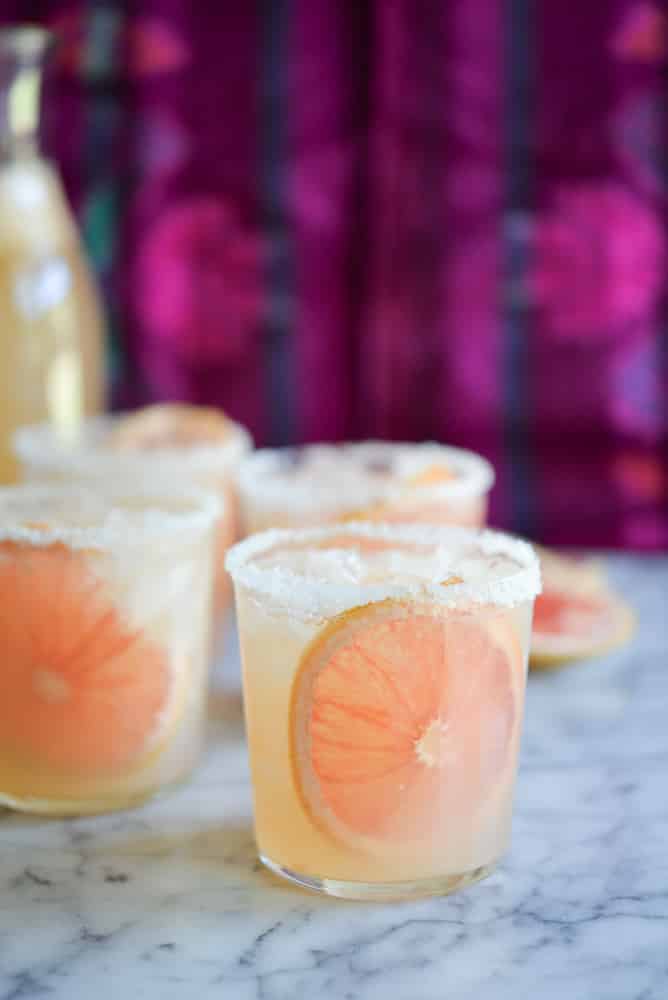 two palomas in glasses with a grapefruit slice and salted rim against a purple background