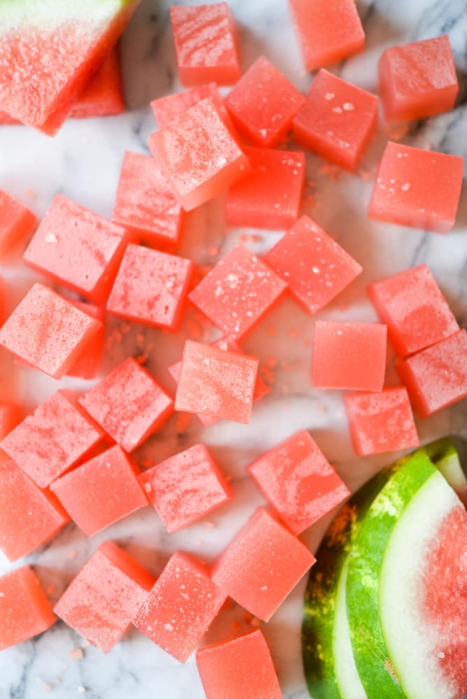 cubed watermelon jello shots on a marble surface with two slices of watermelon