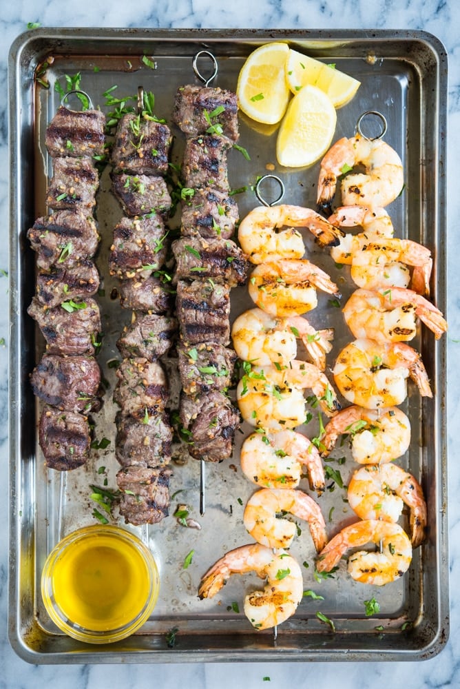 skewers of grilled beef and grilled shrimp on a sheet pan with a bowl of melted butter