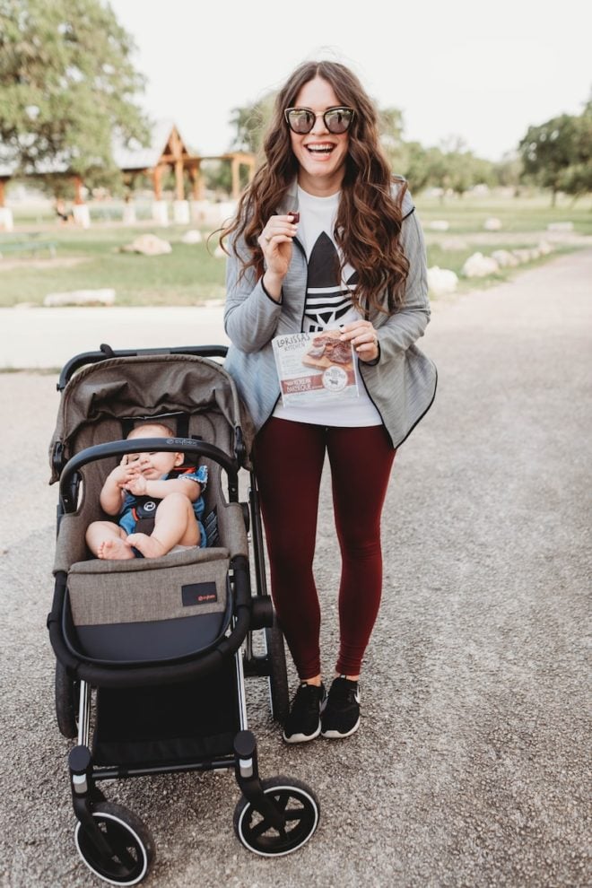 a woman in workout attire with long dark hair holding beef snacks next to a baby in a stroller