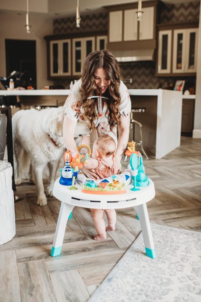 women with long dark hair standing over a baby in an activity chair eating a beef stick with a white dog in the background