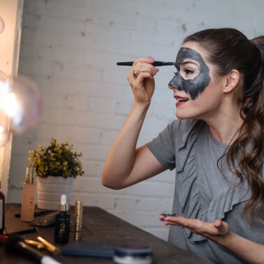 woman putting on a charcoal mask with a brush in front of a lighted mirror - spa day at home