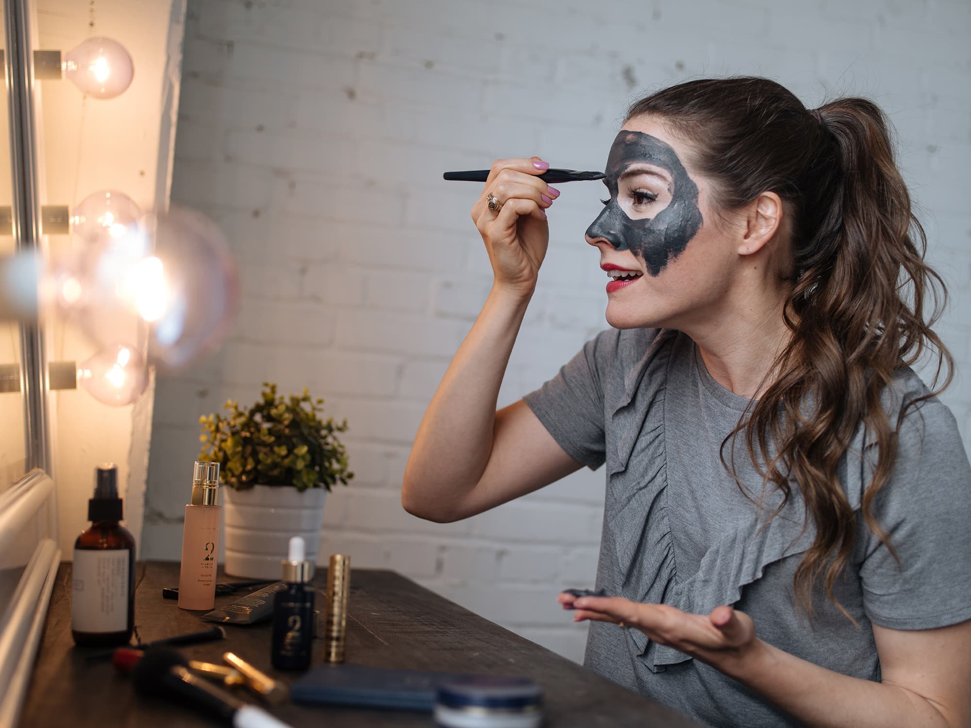 woman putting on a charcoal mask with a brush in front of a lighted mirror - spa day at home