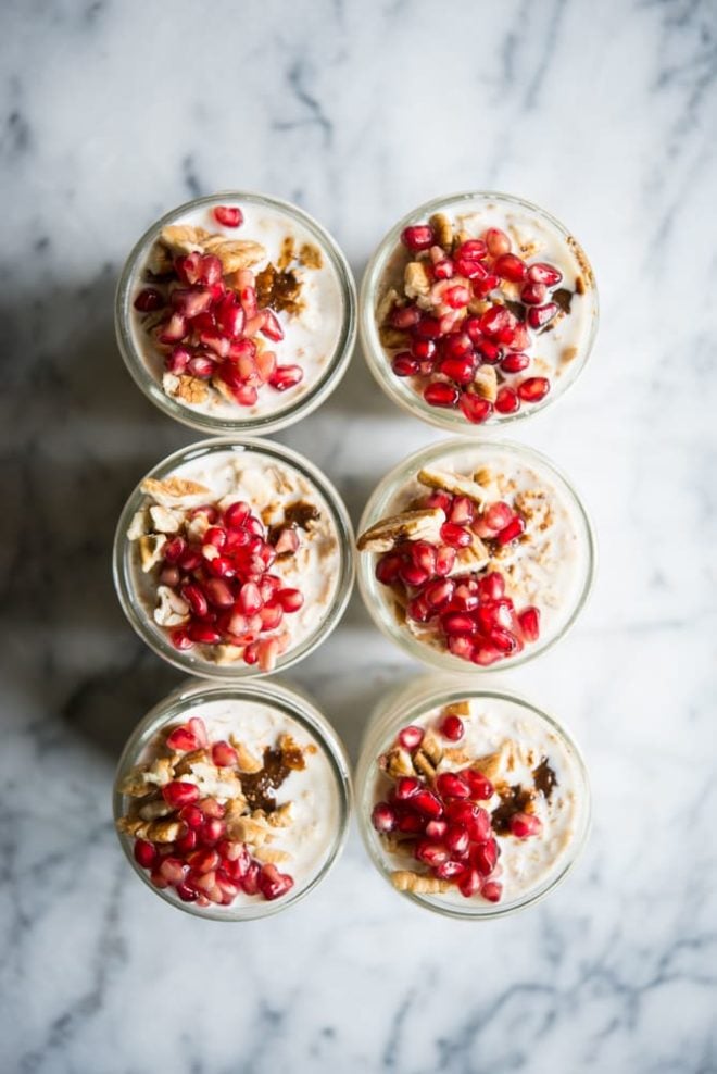 6 glass mason jars filled with overnight oats and topped with pecans and pomegranate seeds on a marble surface