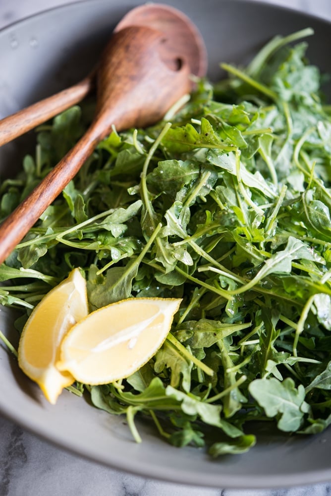 arugula salad in a gray bowl with two lemon wedges and wooden utensils