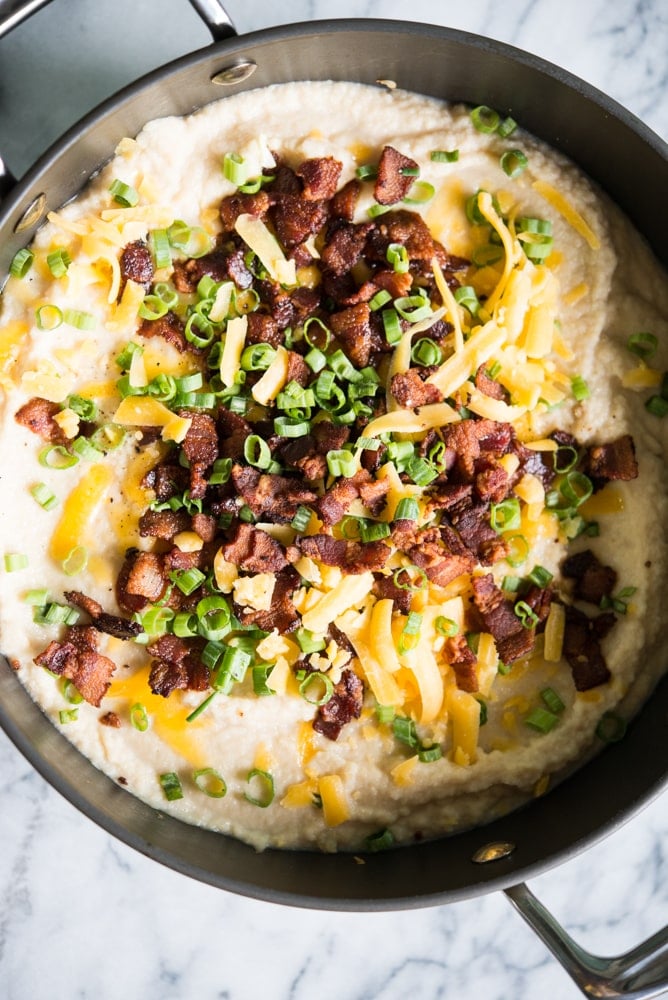 cauliflower soup in a pot topped with cheese, bacon, and green onions on a marble board