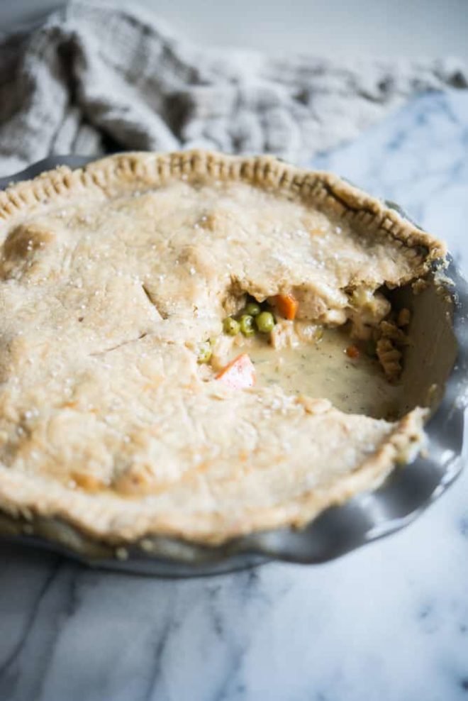 side shot of gluten free chicken pot pie in a grey deep dish pie pan with a scoop removed on a marble surface with a tan towel behind it