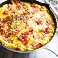 side view of loaded twice baked potatoess topped with melted cheese and crispy bacon in a white enameled cast iron pan on a marble ssurface with a grey towel beside it