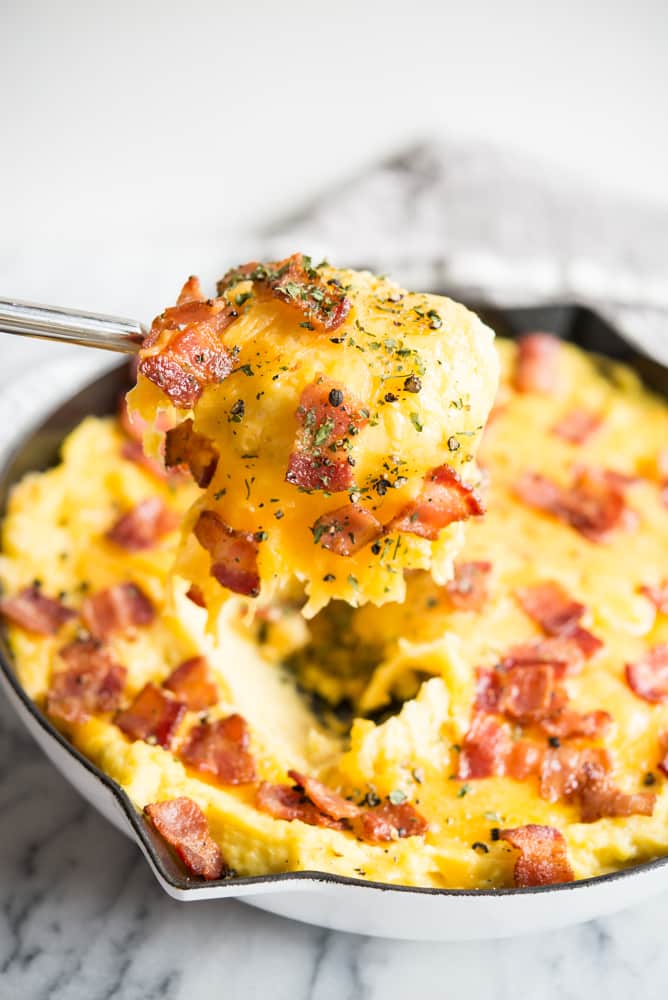 metal spoon holding a spoonful of loaded twice baked potato casserole over a white ceramic cast iron pan full of potato casserole topped with cheese and bacon