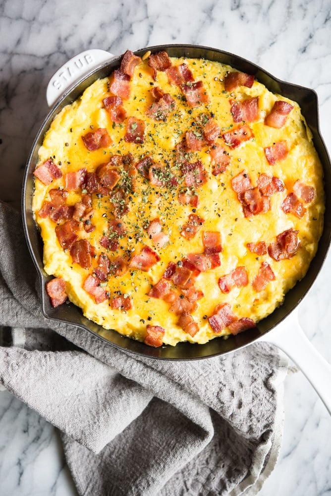 loaded twice baked potatoess topped with melted cheese and crispy bacon in a white enameled cast iron pan on a marble ssurface with a grey towel beside it