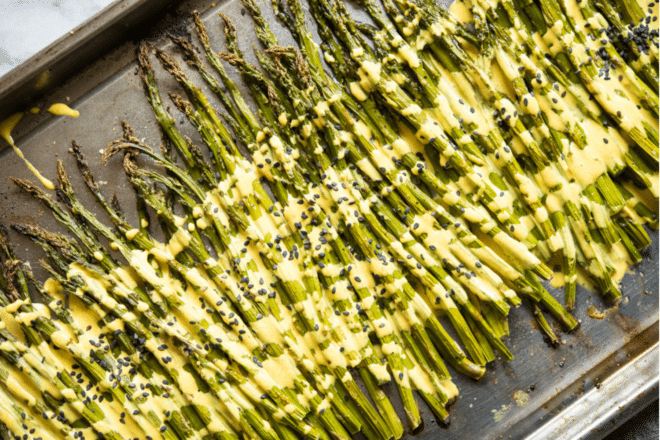 roasted asparagus on a stainless steel sheet pan with a bright yellow lemon cardamom sauce poured over top and garnished with black sesame seeds