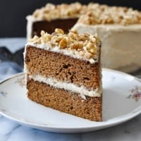 a slice of two layer paleo carrot cake with cream cheese frosting on a white floral plate with the whole cake blurred in the background