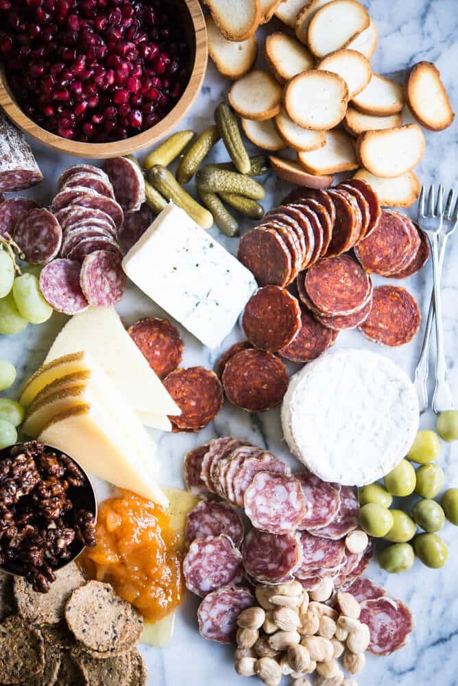 Charcuterie board with meats, cheeses, honey, nuts, a wooden bowl of pomegranates, olives, grapes, pickles, crackers, on a marble board