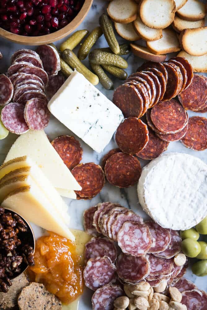 Charcuterie board with meats, cheeses, honey, nuts, a wooden bowl of pomegranates, olives, grapes, pickles, crackers, on a marble board
