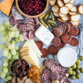 Charcuterie board with meats, cheeses, honey, nuts, a wooden bowl of pomegranates, olives, grapes, pickles, crackers, on a marble board