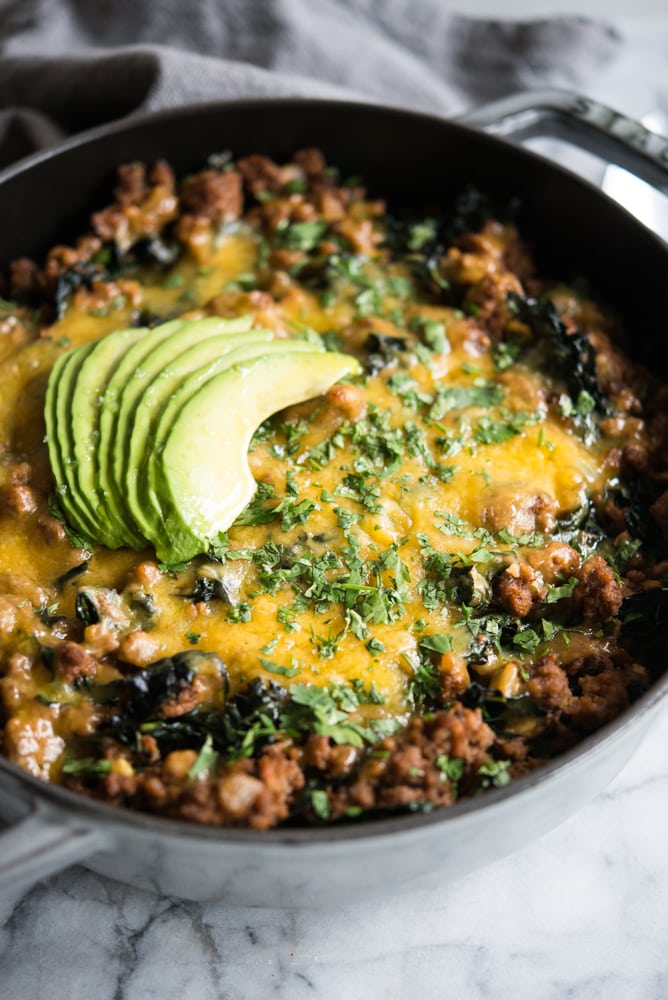 beef taco casserole in a grey cast iron dish topped with melted cheese, chopped cilantro, and sliced avocado