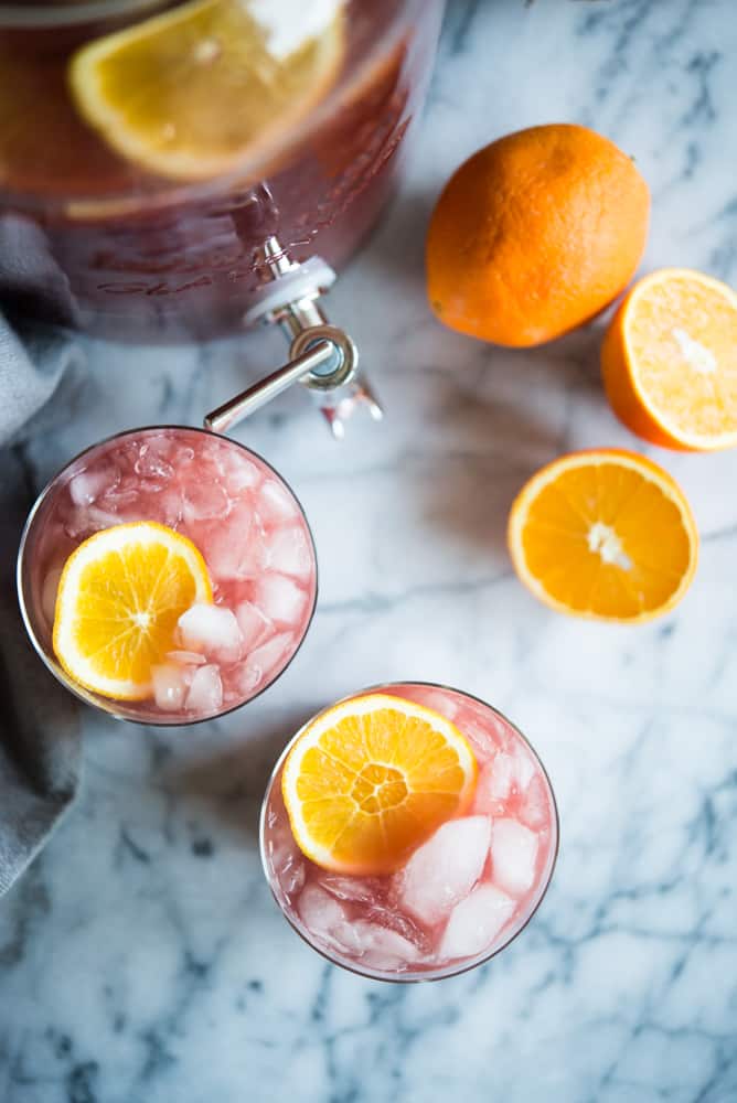 two cups of trash can punch with orange slices in it next to a pitcher and two orange halves, on a marble surface