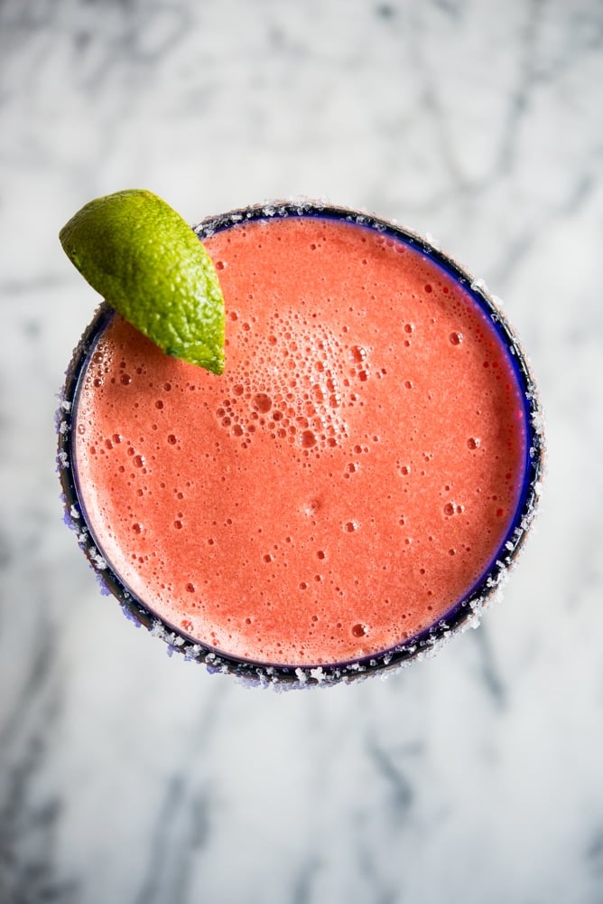 Bright pink frozen watermelon margarita in a blue-rimmed margarita glass with a lime wedge on a marble countertop.