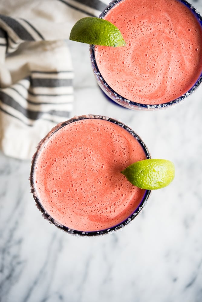Bright pink frozen watermelon margarita in a blue-rimmed margarita glass with a lime wedge on a marble countertop.