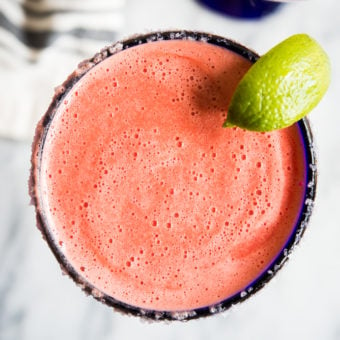 Bright pink frozen watermelon margarita in a blue-rimmed margarita glass with a lime wedge on a marble countertop.