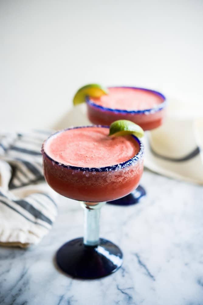 frozen watermelon margaritas in a blue-rimmed margarita glass with a salted rim and lime wedge on a marble surface