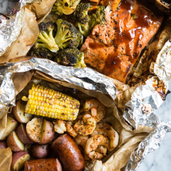 2 grilled foil packet dinners - shrimp, sausage, potatoes, and corn with cajun seasoning in a parchment and foil package and bbq salmon, sweet potatoes, and broccoli in a foil packet.