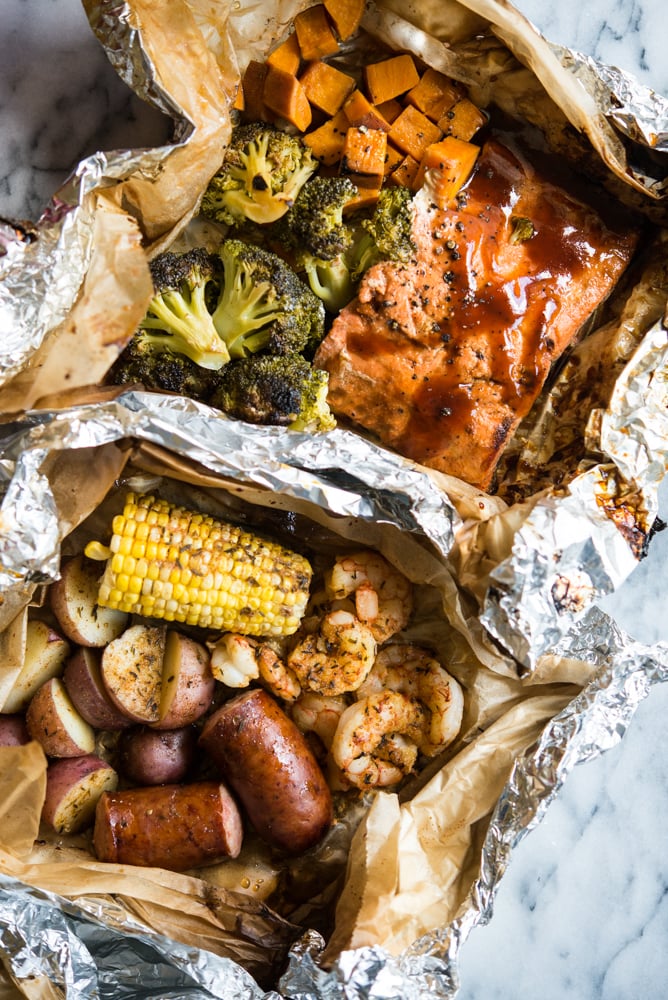 2 grilled foil packet dinners - shrimp, sausage, potatoes, and corn with cajun seasoning in a parchment and foil package and bbq salmon, sweet potatoes, and broccoli in a foil packet.