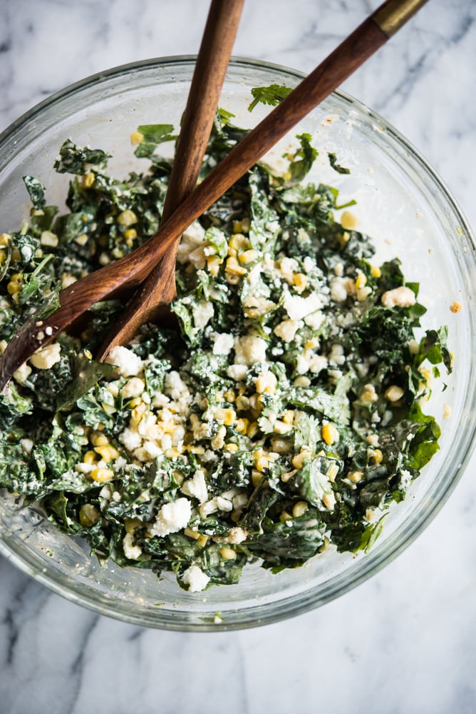 kale slaw in a bowl with corn and crumbled cotija cheese in a glass bowl with wooden serving spoons on a marble board