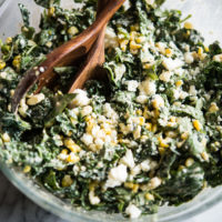 kale slaw in a bowl with corn and crumbled cotija cheese in a glass bowl with wooden serving spoons on a marble board