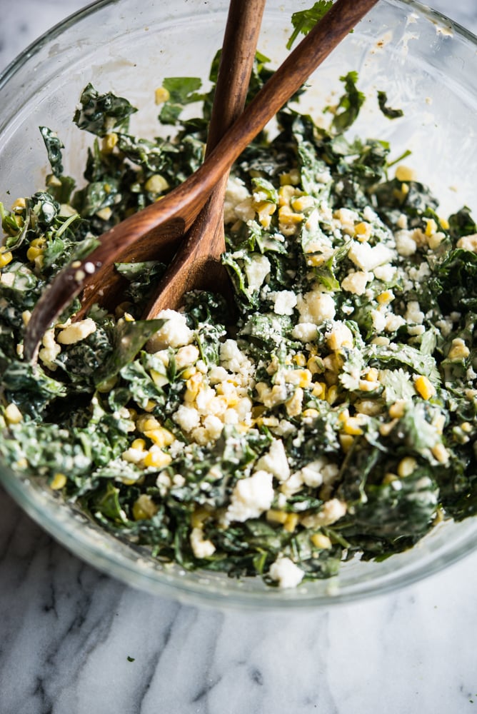 kale slaw in a bowl with corn and crumbled cotija cheese in a glass bowl with wooden serving spoons on a marble board