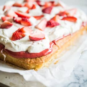 Strawberry shortcake - Vanilla sheet cake topped with whipped cream and strawberries on parchment paper on a marble slab.