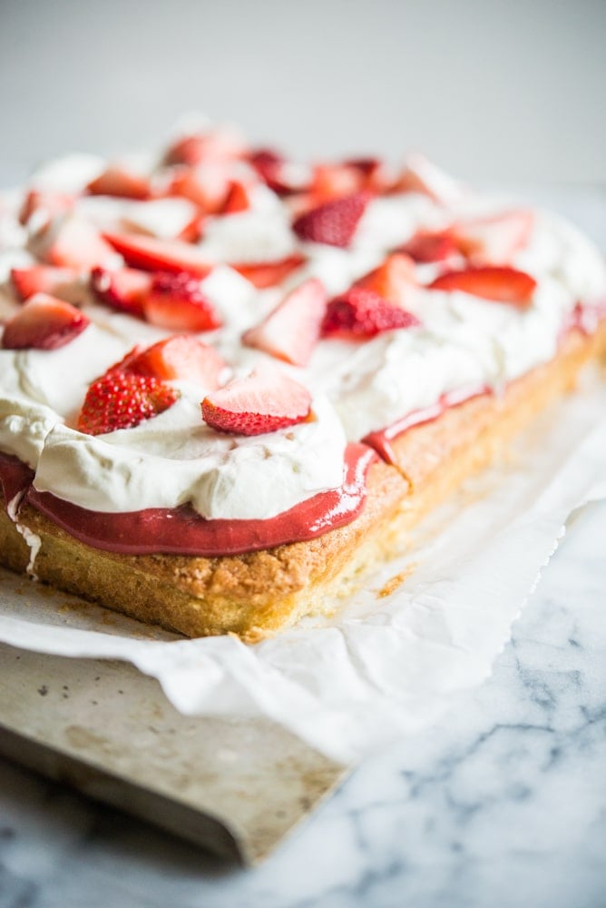 Strawberry shortcake - Vanilla sheet cake topped with whipped cream and strawberries on parchment paper on a marble slab.