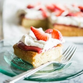 Strawberry shortcake - a square of vanilla cake is topped with strawberry sauce, whipped cream, and fresh strawberries and sits on a clear glass plate with a fork on a marble countertop.