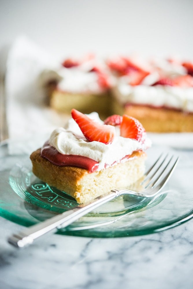 Strawberry shortcake - a square of vanilla cake is topped with strawberry sauce, whipped cream, and fresh strawberries and sits on a clear glass plate with a fork on a marble countertop.