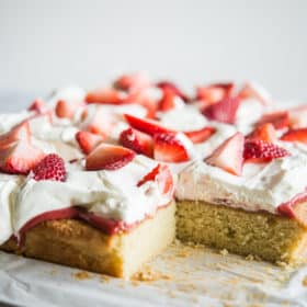 Strawberry shortcake - Vanilla sheet cake topped with whipped cream and strawberries, cut into squares with one slice missing. The cake is place on top of parchment paper on a marble slab.