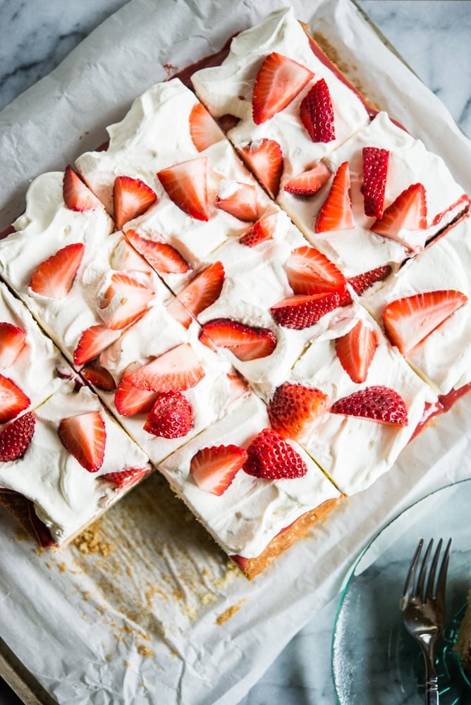 Strawberry shortcake - Vanilla sheet cake topped with whipped cream and strawberries, cut into squares with one slice missing. The cake is place on top of parchment paper on a marble slab.