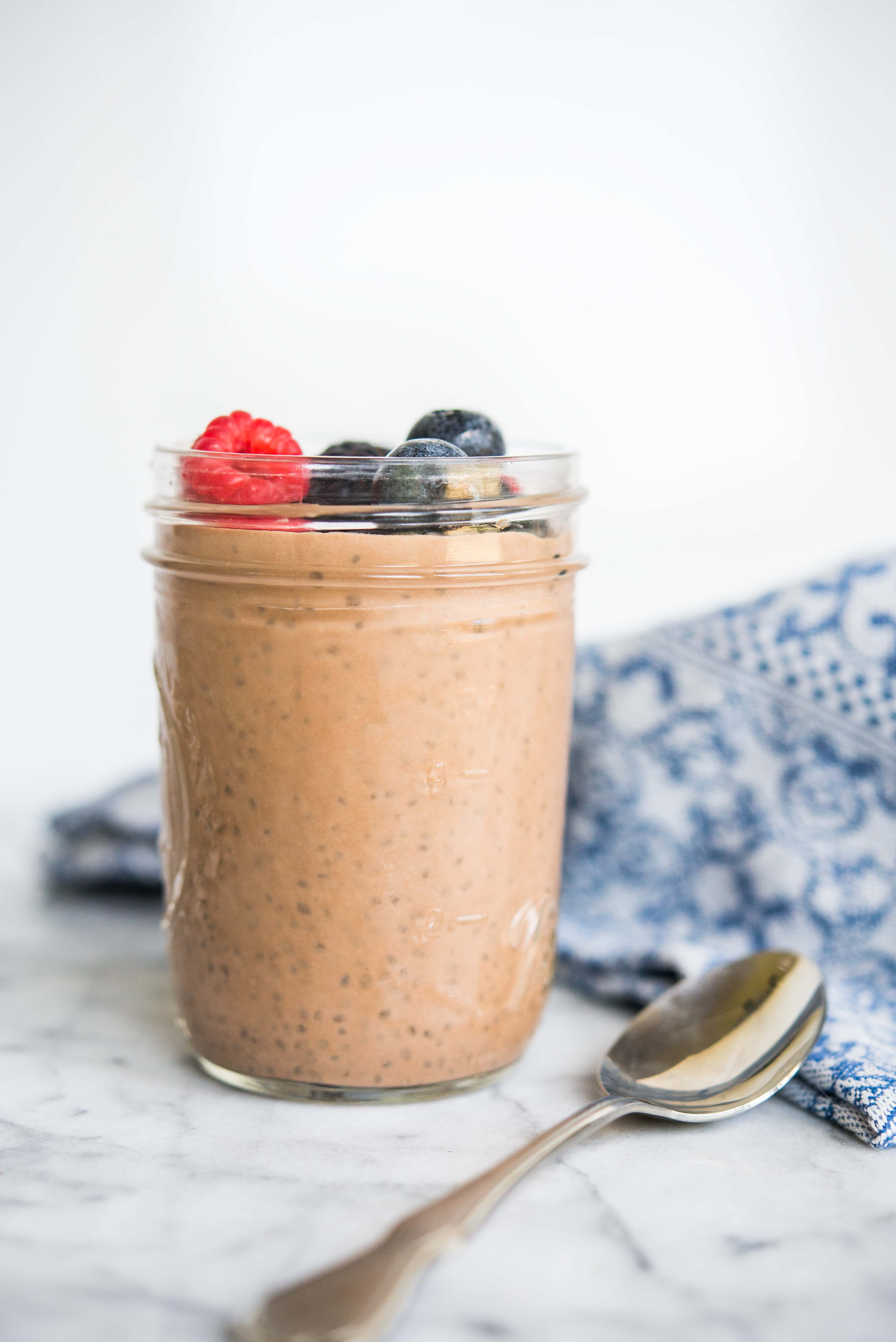 chocolate chia pudding in a mason jar topped with berries on a marble board with a spoon and blue towel