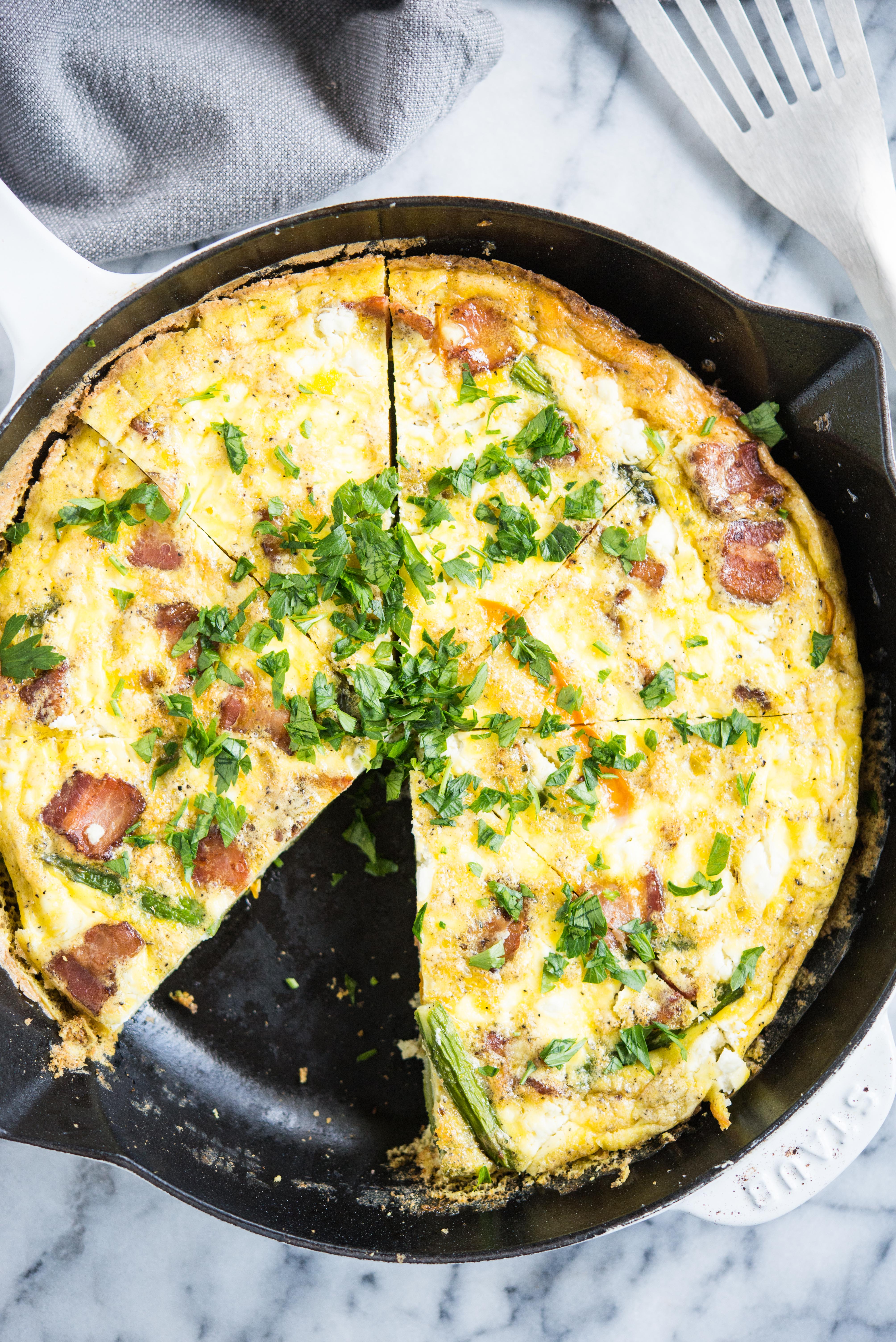 frittata with asparagus, goat cheese, and bacon in a cast iron pan with one slice missing on a marble board