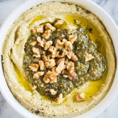 creamy hummus with pesto and walnuts on top in a white bowl on a marble surface