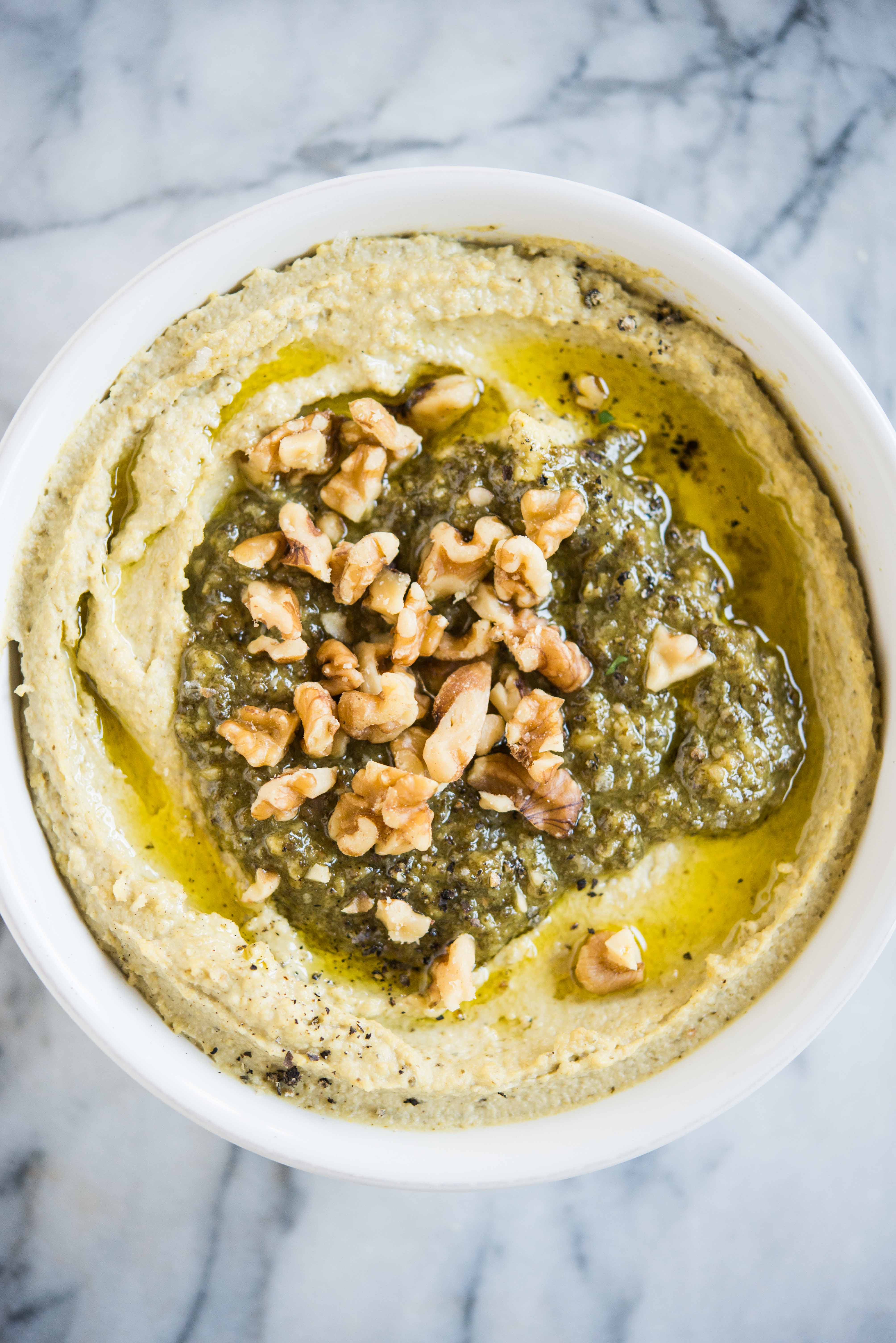 creamy hummus with pesto and walnuts on top in a white bowl on a marble surface