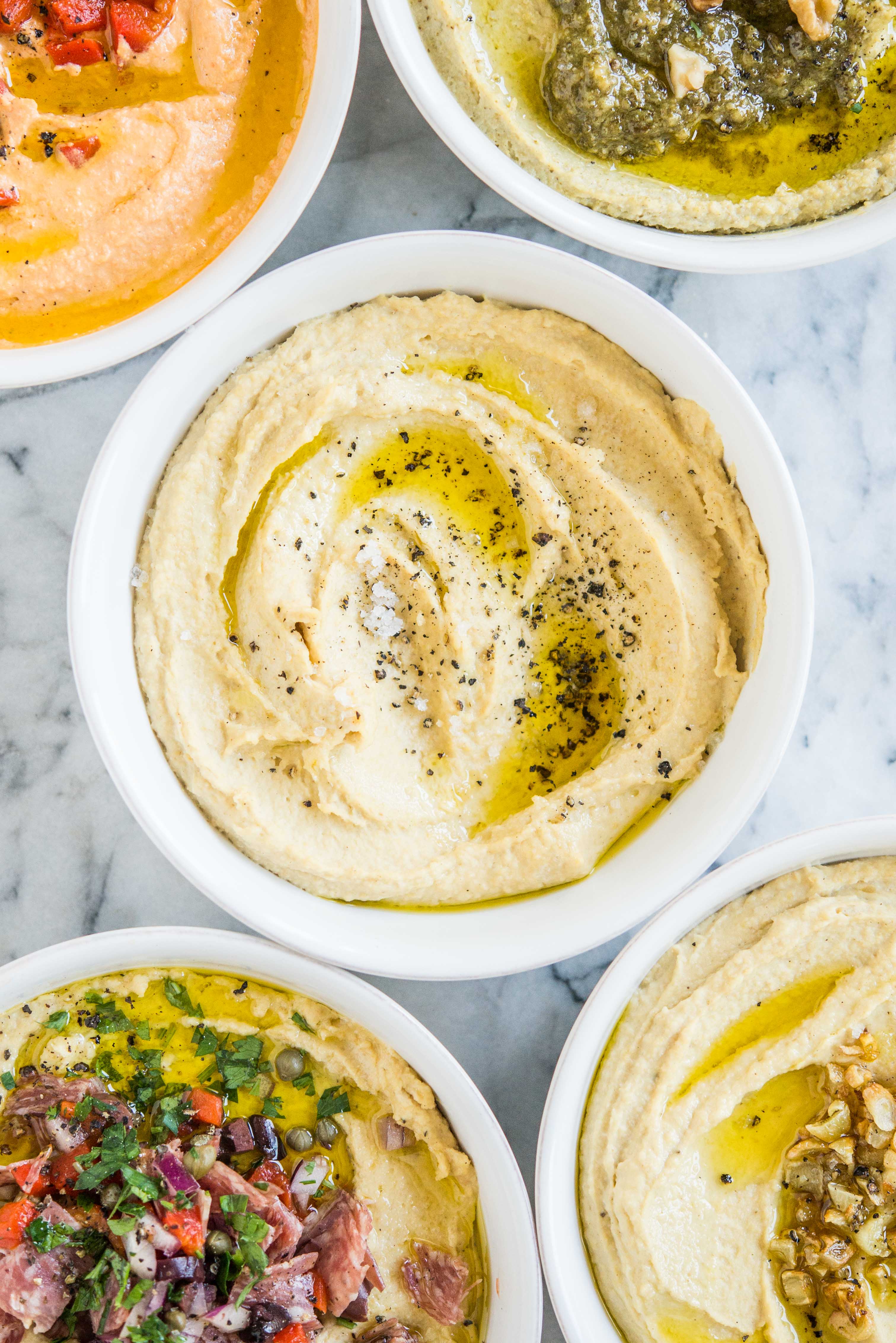 five bowls of hummus with one bowl of plain hummus in the center sprinkled with black pepper