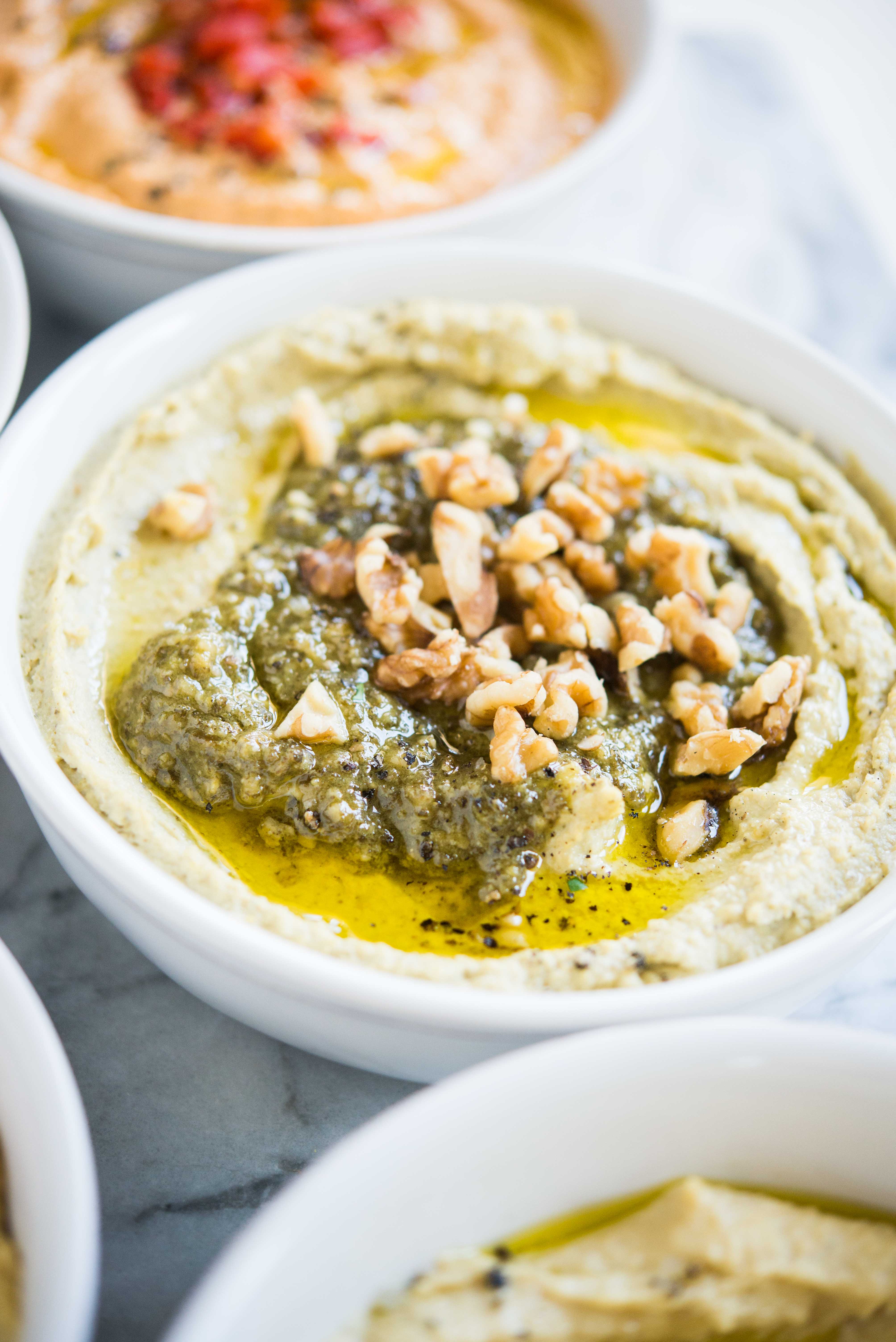 creamy hummus with pesto and walnuts on top in a white bowl on a marble surface