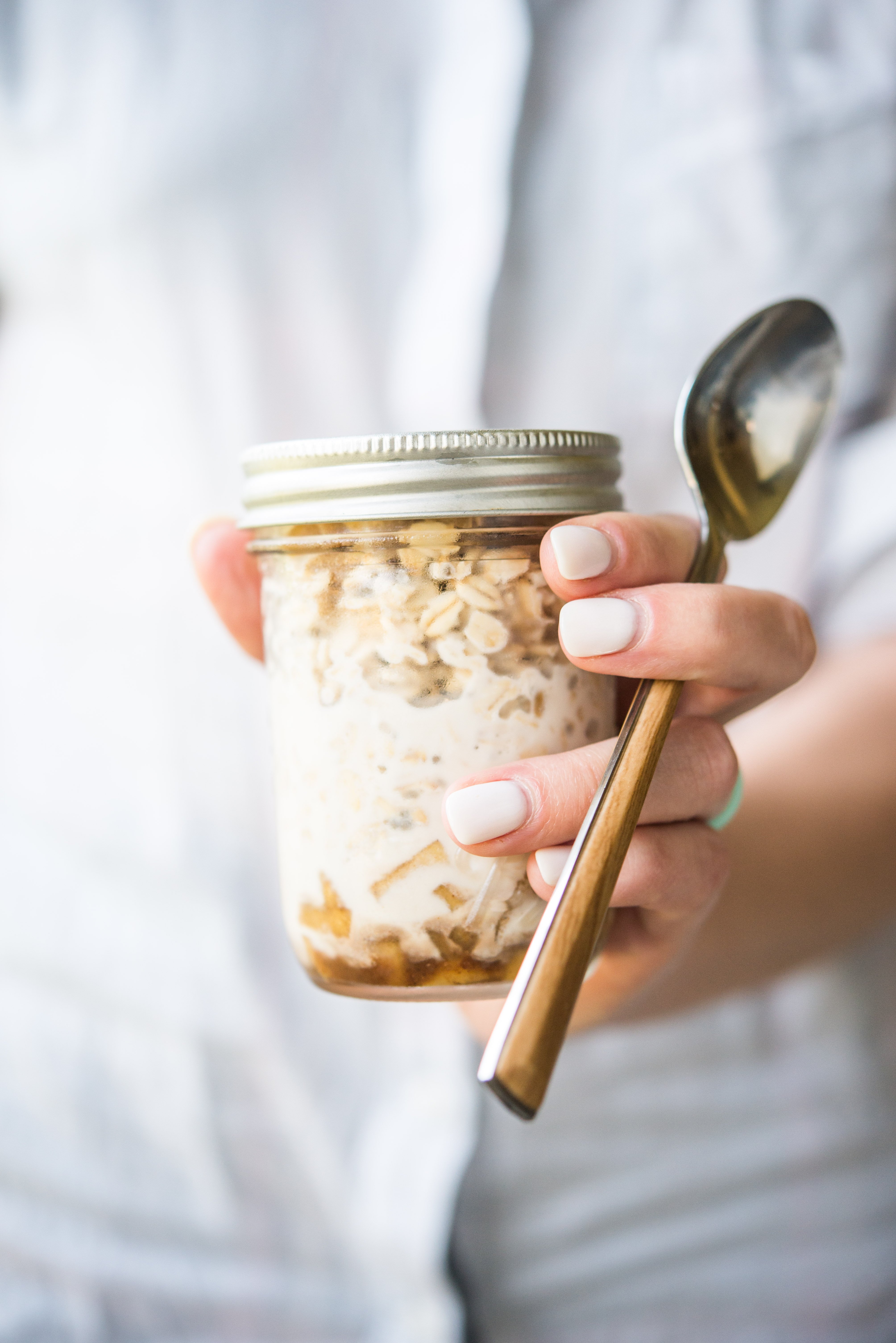 woman in a white shirt holding a mason jar filled with apple cinnamon overnight oats and a spoon